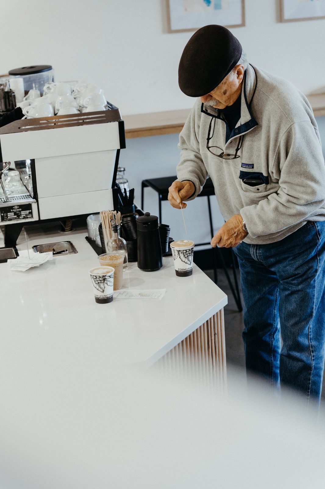 Older man in a flat cap stirring his latte at the pickup counter