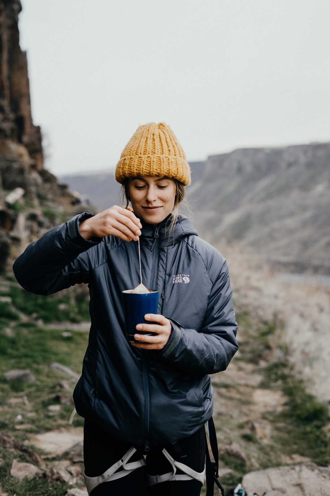 Woman in a yellow beanie dunking a tea bag into a blue cup at a canyon overlook