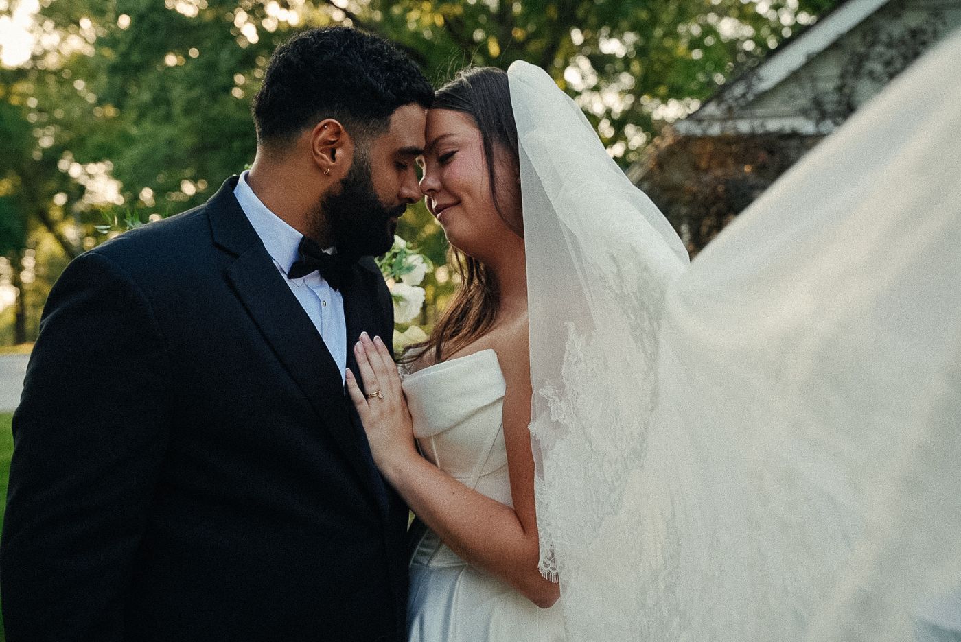 A groom in a black tuxedo and bow tie touching foreheads with his emotional bride as her long lace veil blows in the wind