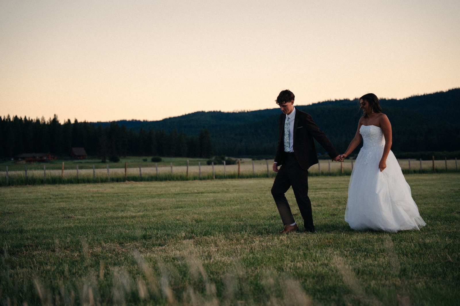 A bride and groom holding hands and walking through a grassy field at sunset, with pine-covered hills behind them