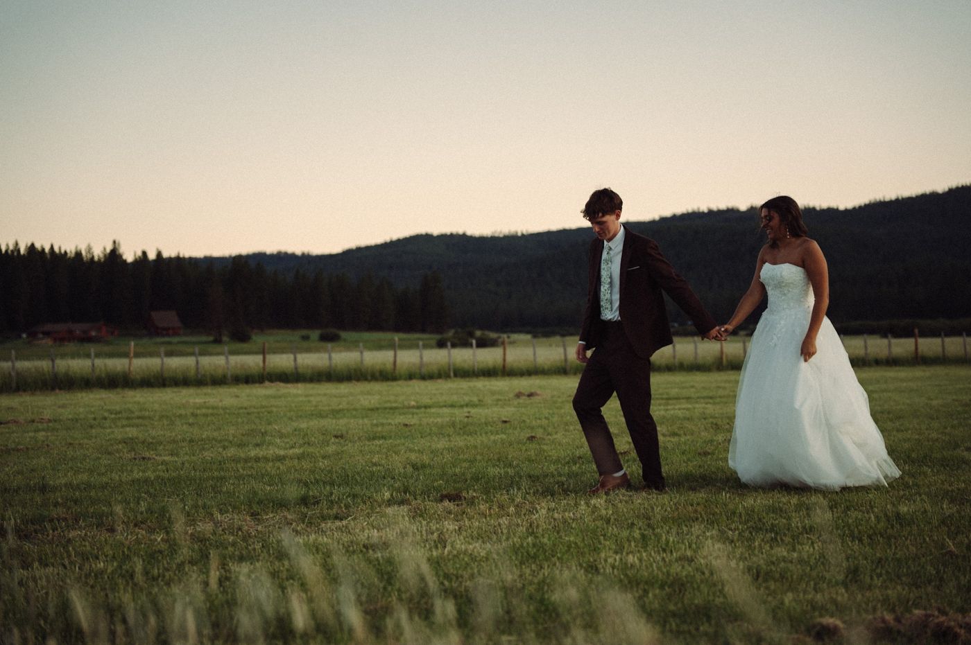 A bride in a white gown and groom in a dark suit walking hand in hand through a meadow at dusk, with forested hills behind them