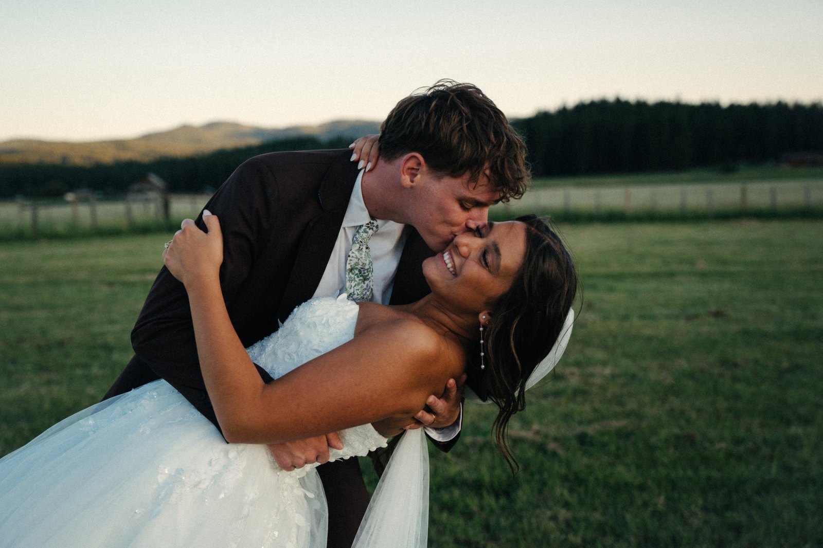 A groom in a dark suit dipping his laughing bride and kissing her cheek in a grassy Idaho field at sunset, with hills and pine trees behind them