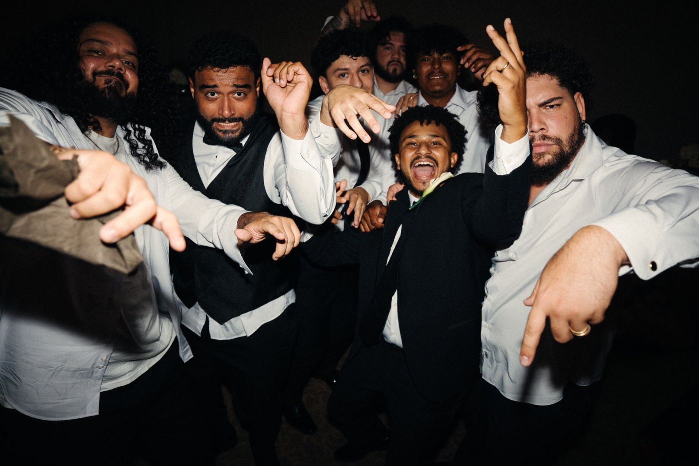 A groom in a black suit and his groomsmen in white shirts cheering and throwing hand signs at the camera on the dance floor