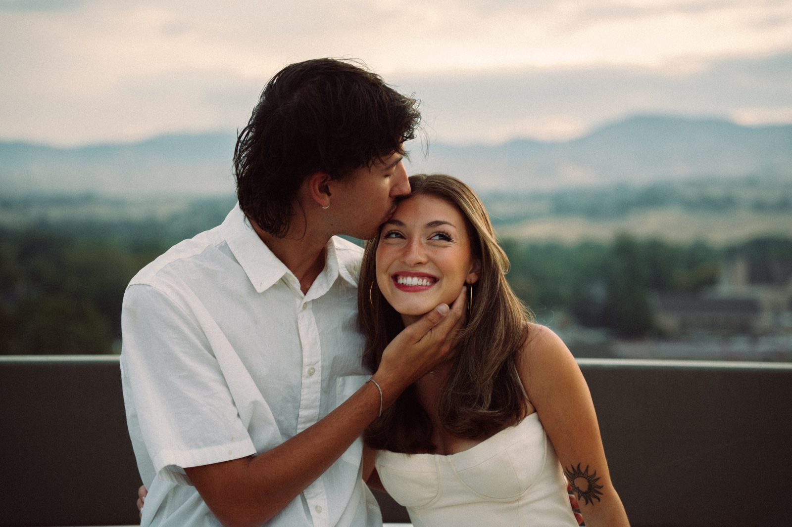 Lucas kisses Audry's forehead as she smiles up at him on a Boise rooftop with the foothills behind them