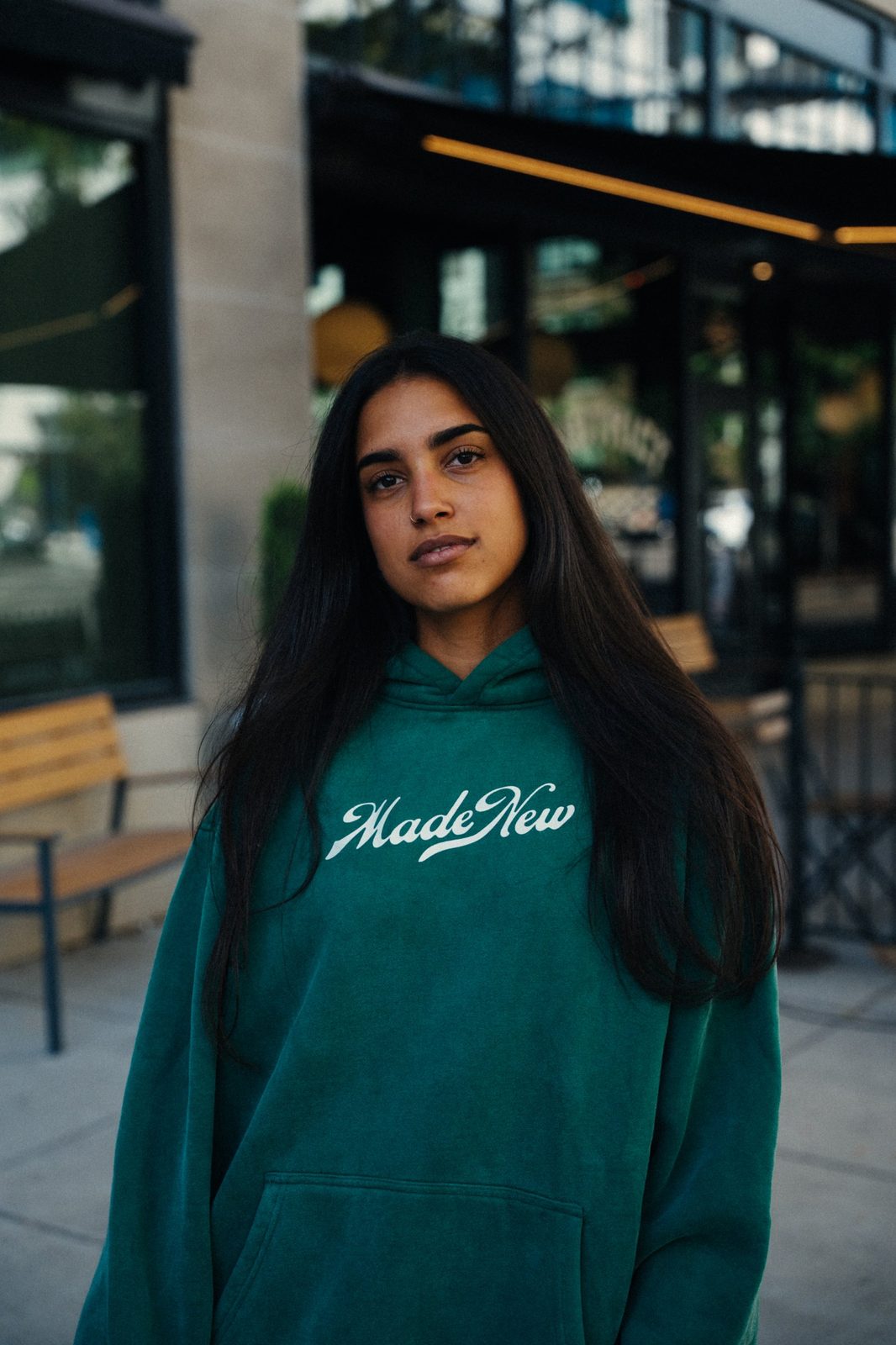 Woman with long dark hair looking at the camera in a green Made New hoodie on a city sidewalk outside a cafe