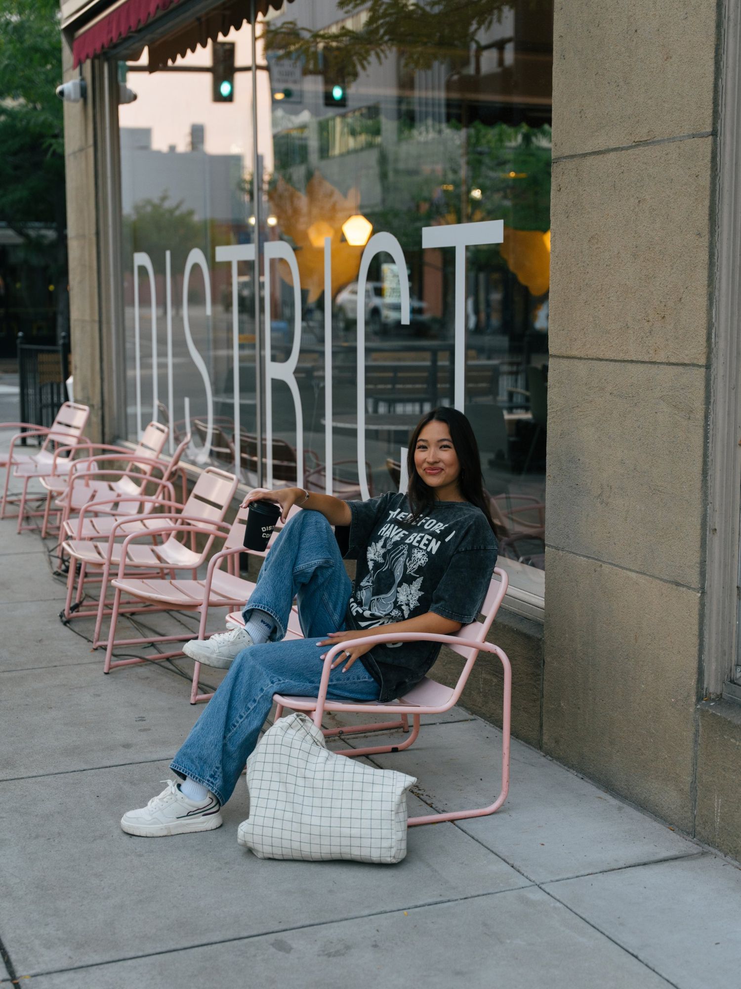 Woman in a Made New graphic tee and wide-leg jeans, seated in a pink chair outside a coffee shop window with a coffee cup in her hand