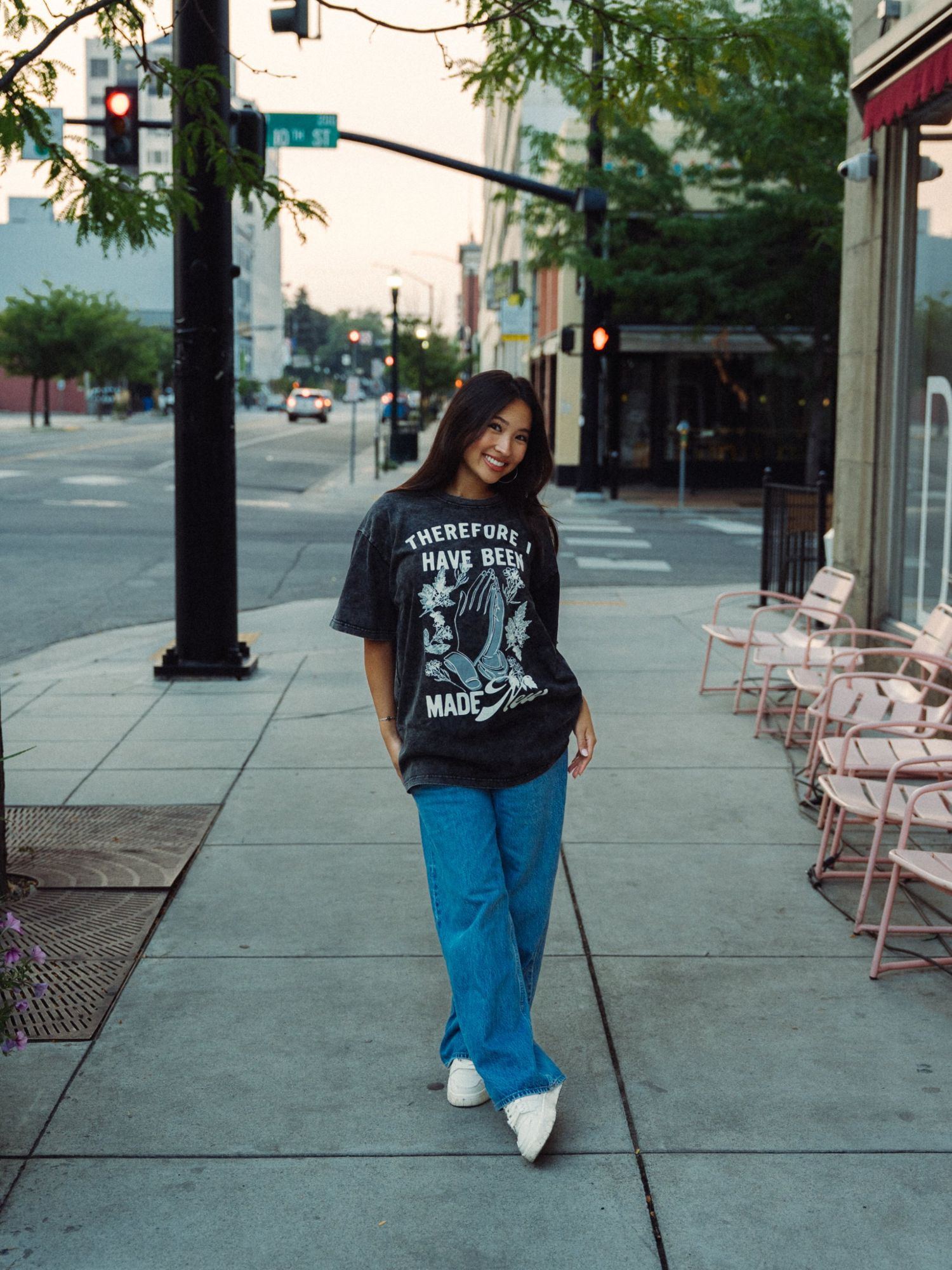 Woman smiling and tilting her head on a downtown sidewalk, in a washed black Therefore I have been made new tee and blue jeans