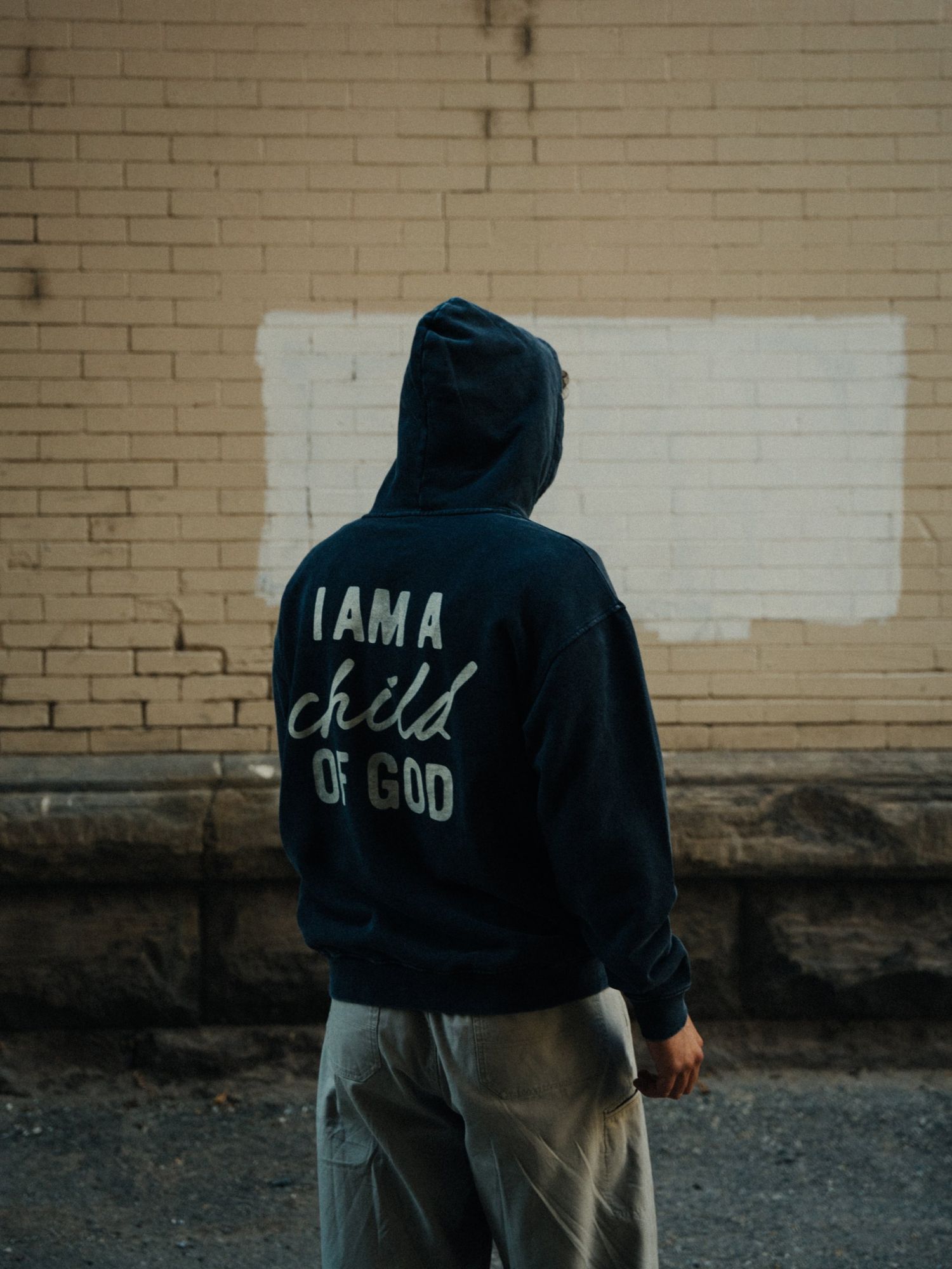 Person seen from behind in a navy hooded sweatshirt that reads I am a child of God, standing in front of a cream brick wall with a painted white patch