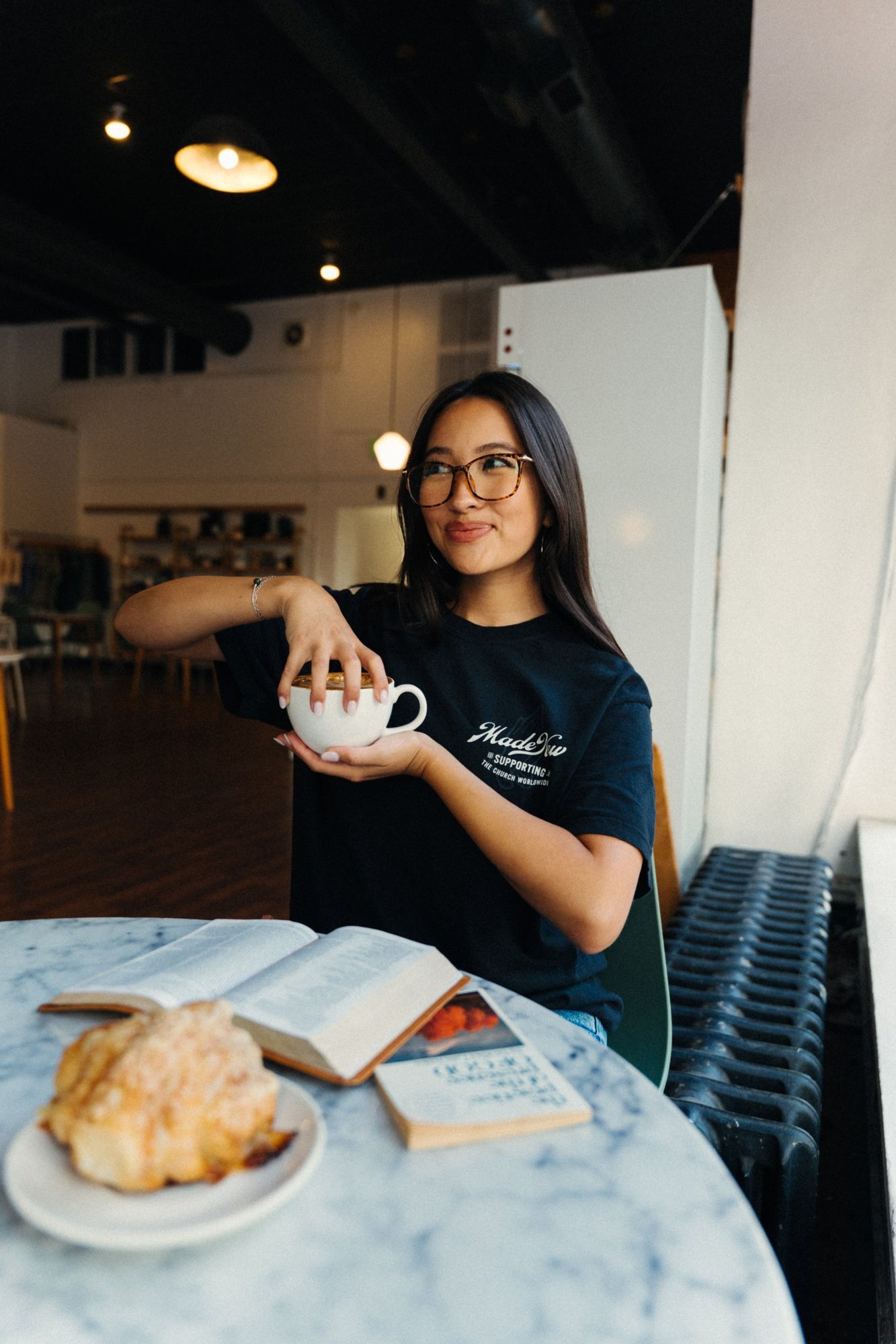 Woman in glasses and a navy Made New tee laughing off camera, holding a latte at a marble table with a pastry and an open Bible