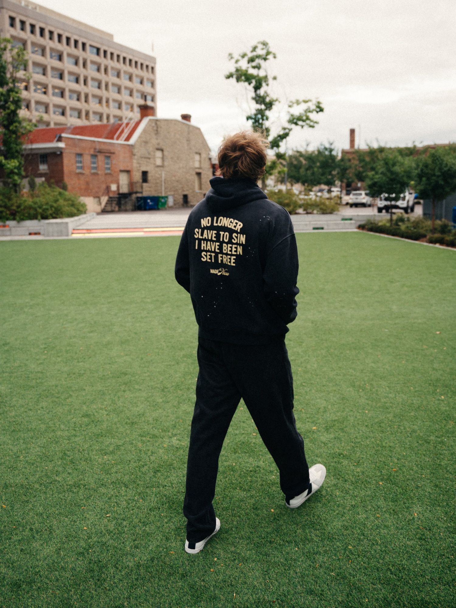 Person walking away across a turf lawn in a black hoodie printed No longer slave to sin, I have been set free, with brick buildings behind