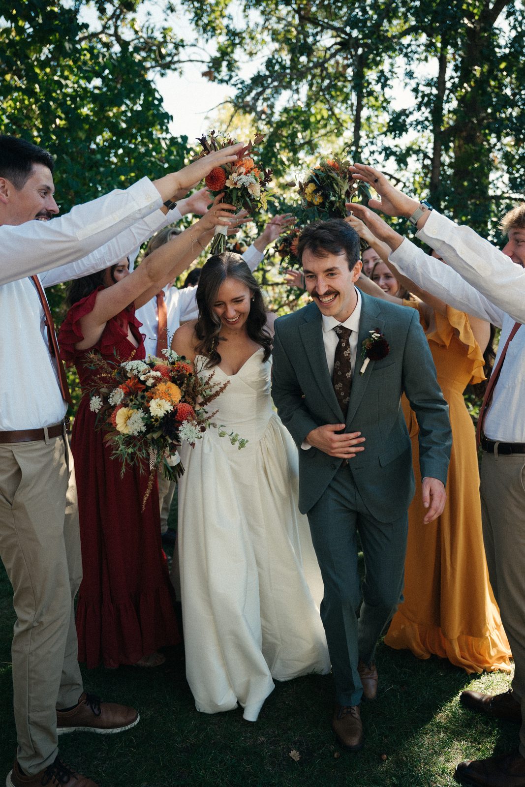 Bride and groom walk laughing under an arch of friends' arms and bouquets