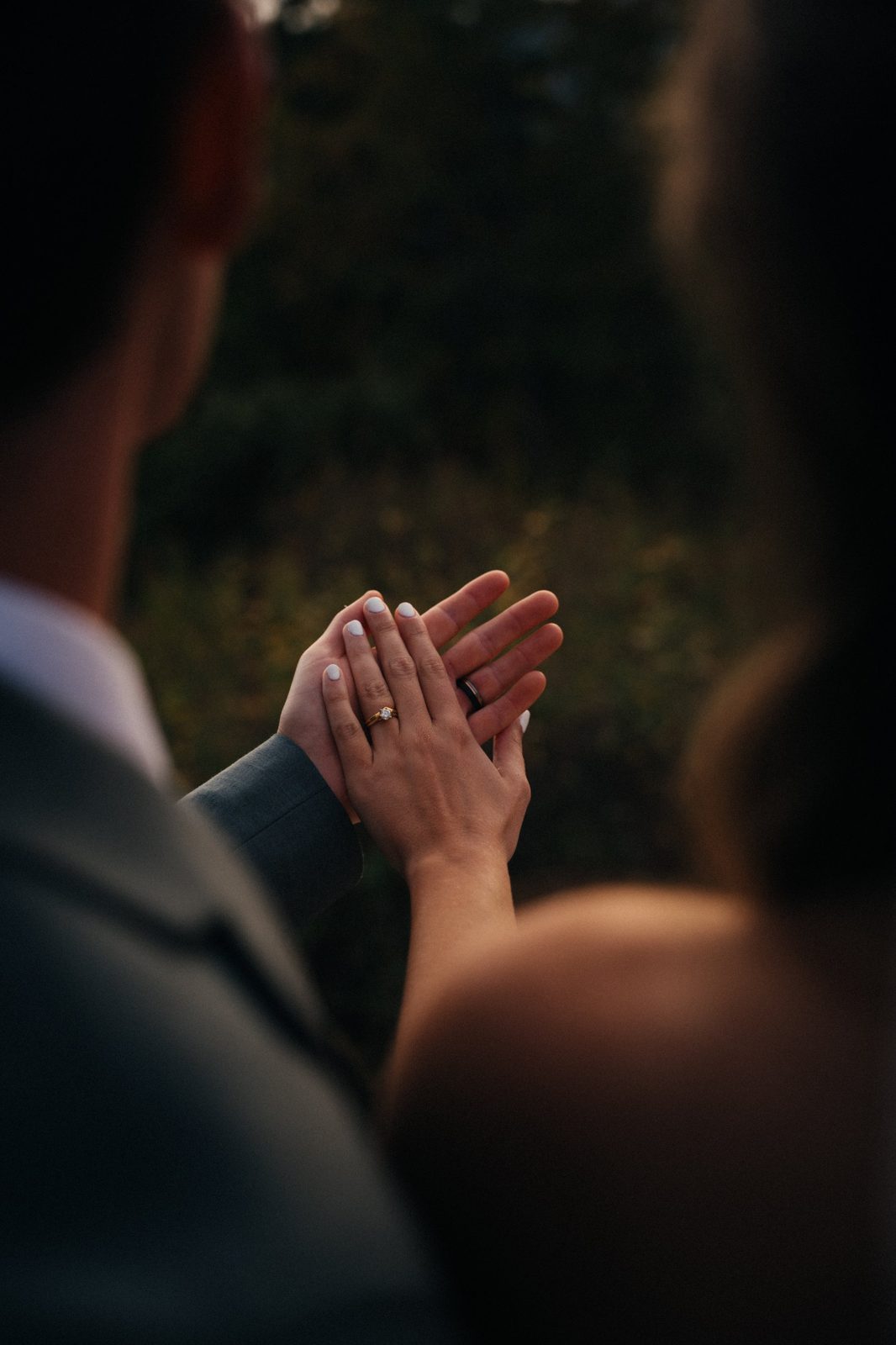 Close-up of a bride and groom's hands together, her engagement ring and his black wedding band catching the evening light