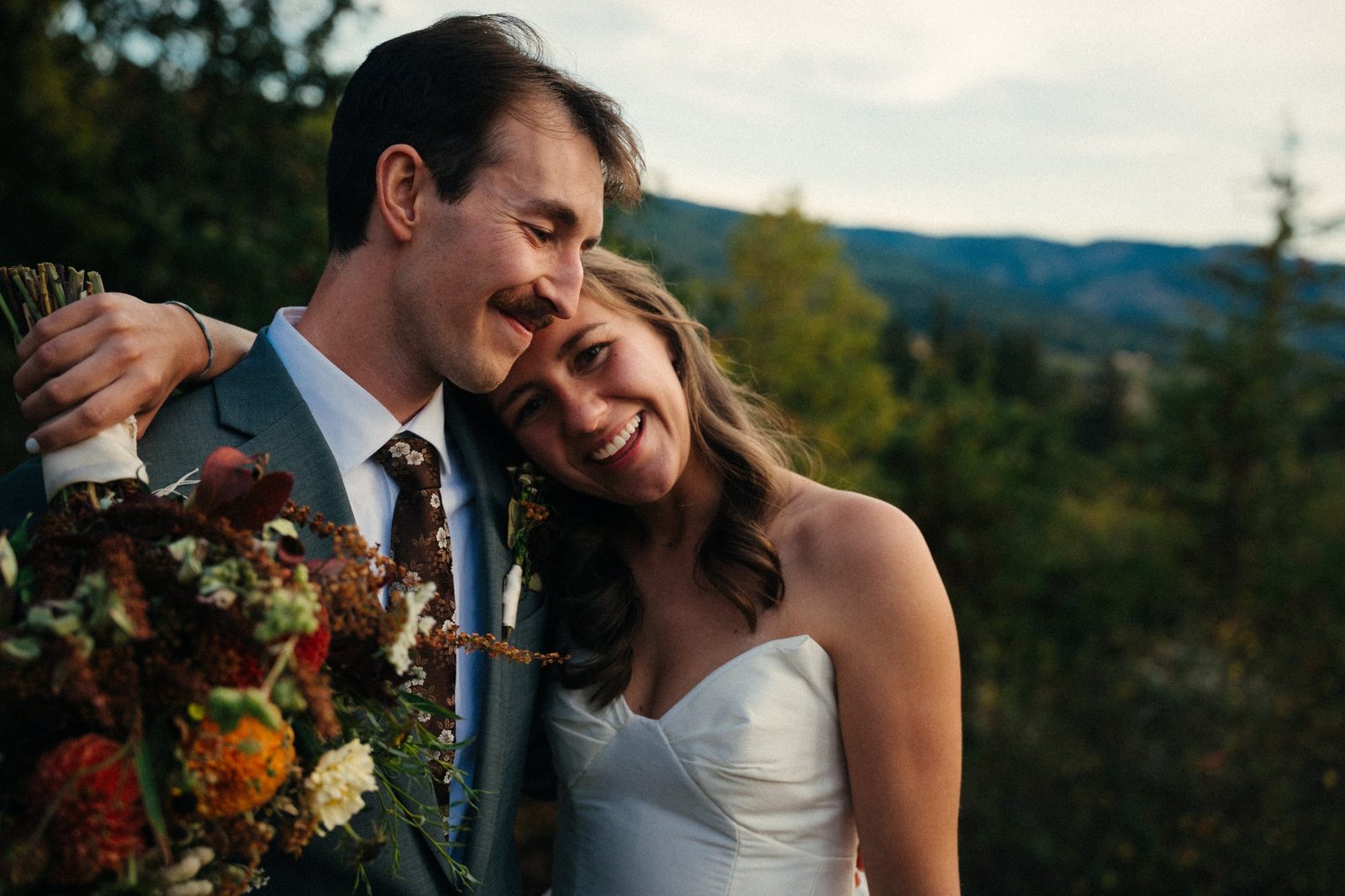 Groom smiling down with his arm around his laughing bride, a fall bouquet in the foreground and hills behind them