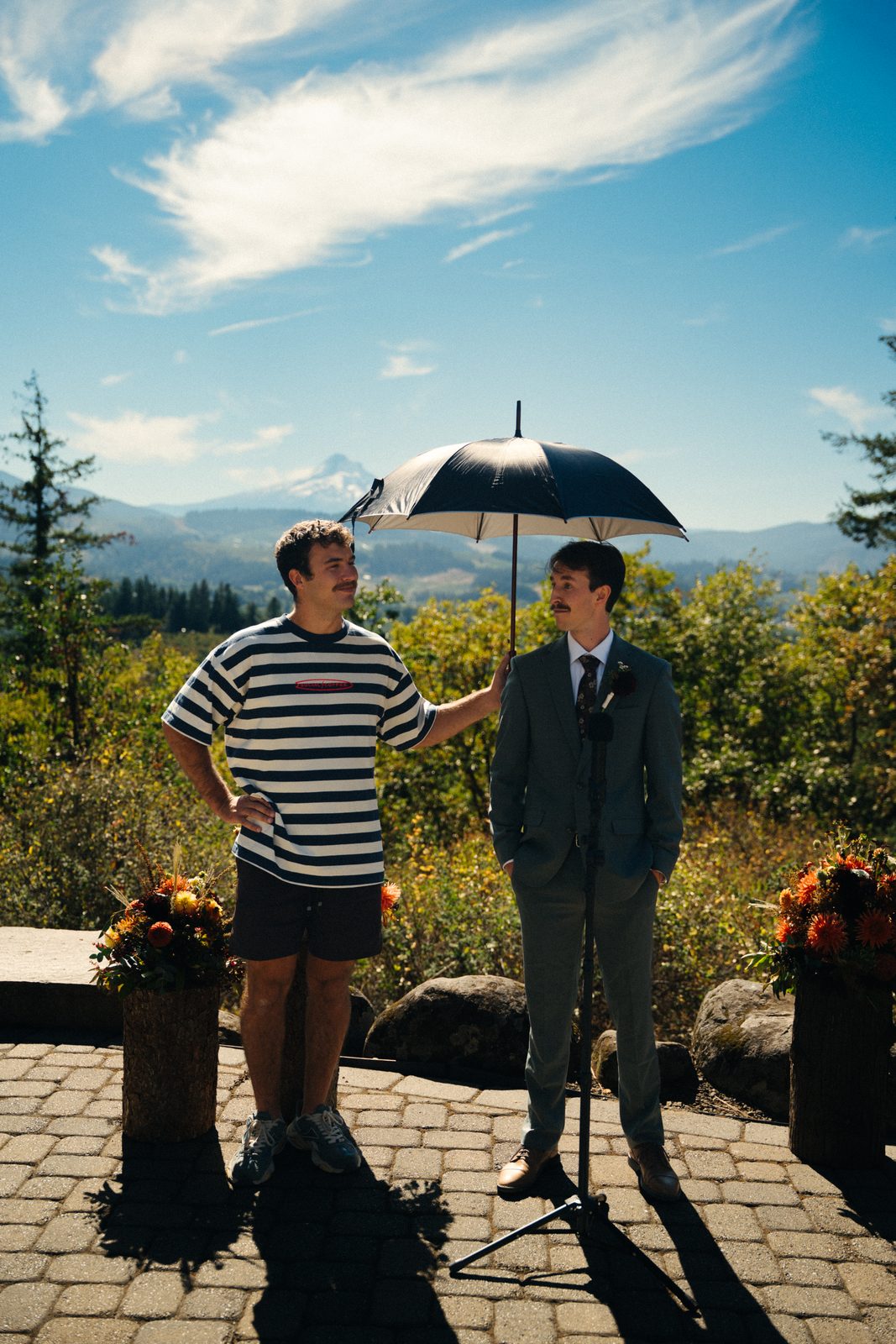 Groom in a green suit standing under an umbrella held by a friend in a striped shirt, Mount Hood behind them