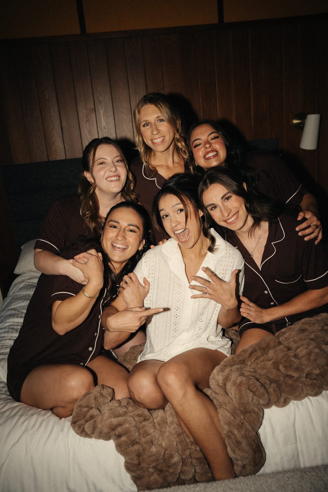 Becca and her five bridesmaids piled together on the bed, laughing, as she shows off her engagement ring