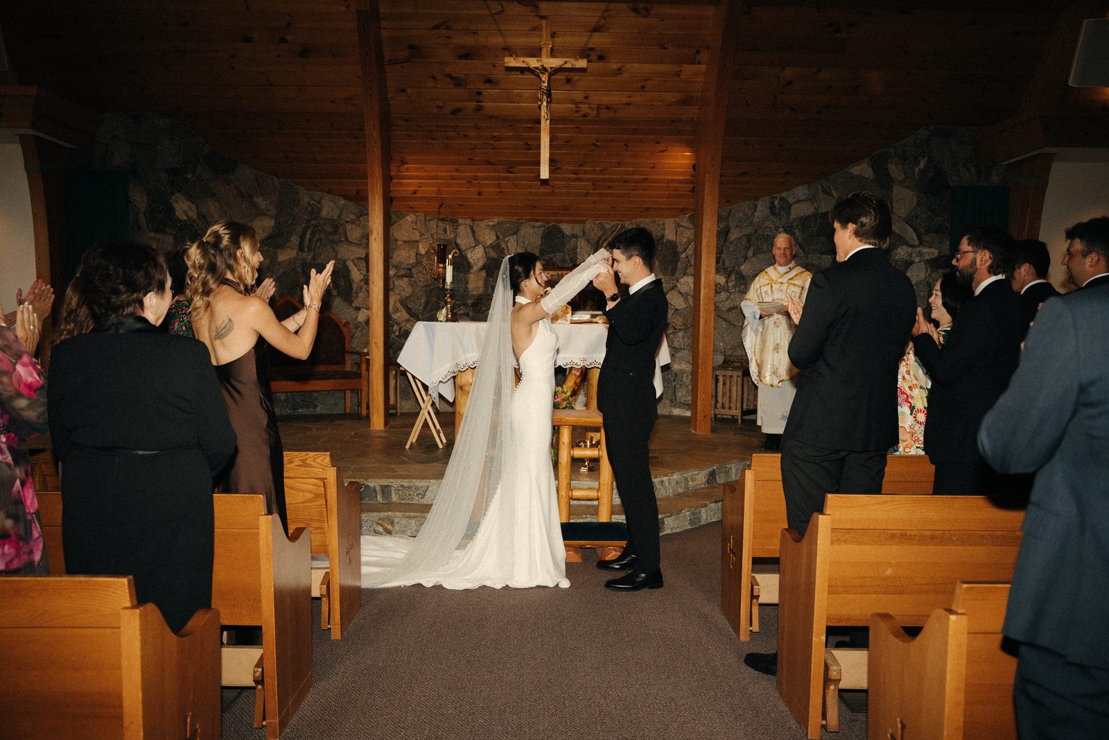 Becca and Skyler raise their joined hands at the altar as guests applaud and the priest looks on