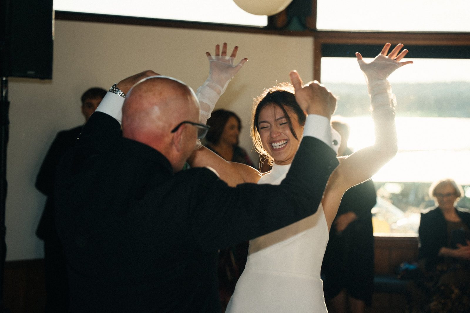 Becca laughing with her hands in the air while dancing with her dad, with the lake glowing in the window behind them