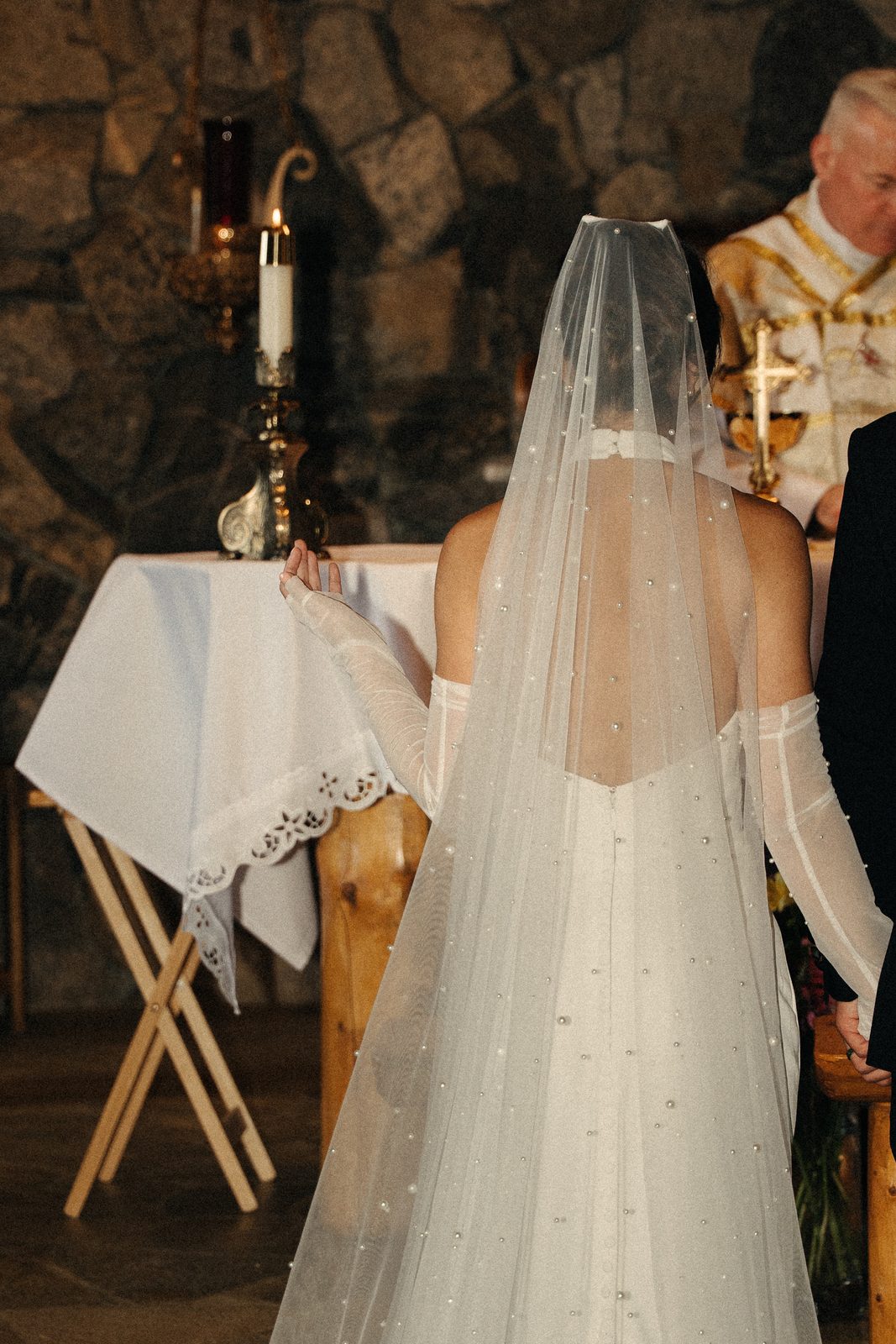 Becca from behind in a pearl-covered veil at the altar of a stone chapel, with a candle lit beside her