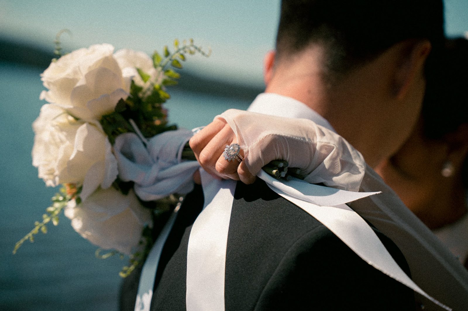 Becca’s ringed hand and white rose bouquet resting on Skyler’s shoulder as they embrace by the lake