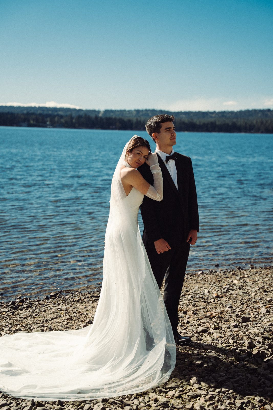 Becca leaning her head on Skyler’s shoulder as he looks out over the water at a lake in McCall, Idaho, her long veil trailing on the rocks