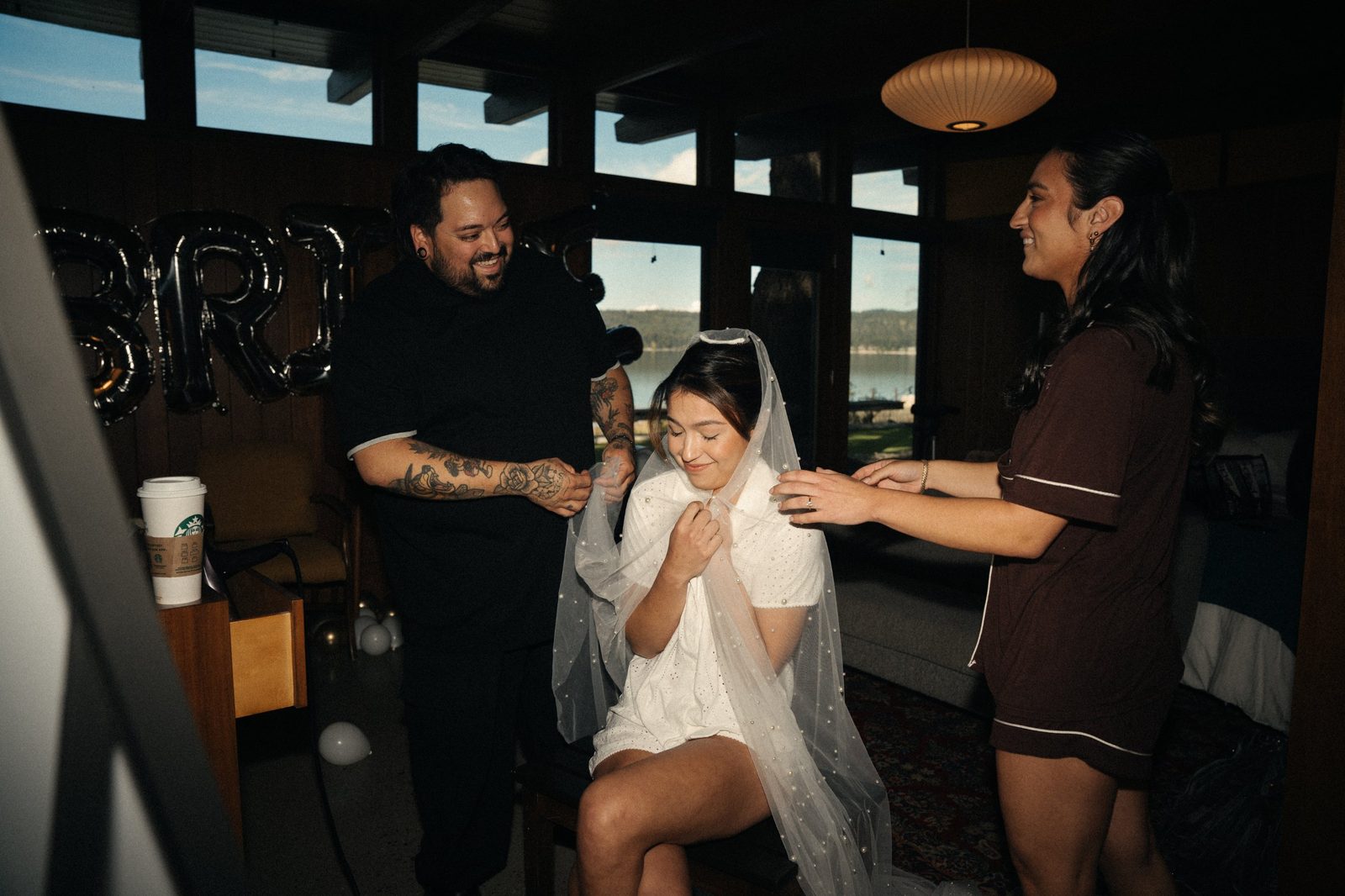 Becca smiling down while a bridesmaid and her stylist help drape a pearl veil over her shoulders