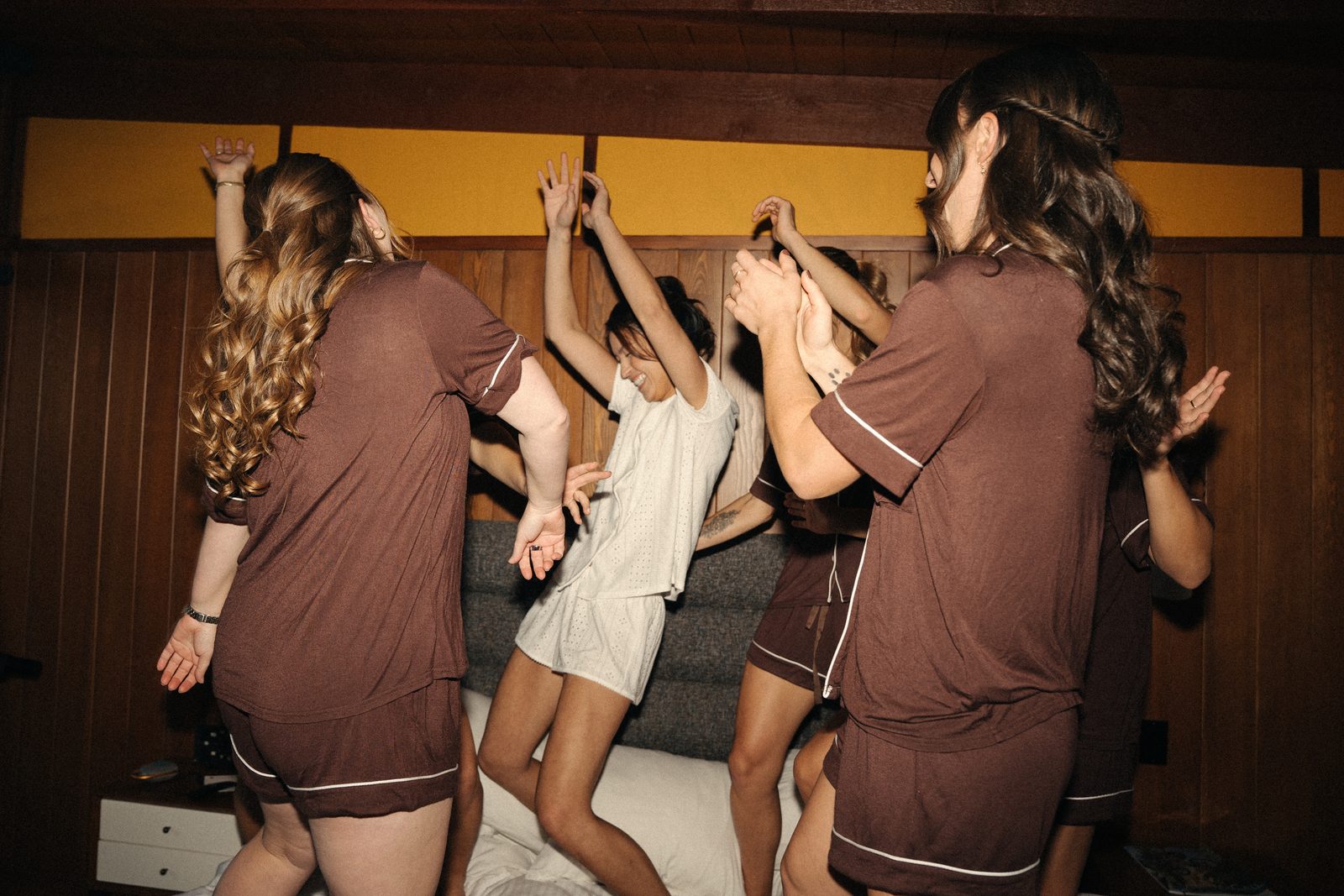 Becca dancing with her arms up on the bed while her bridesmaids in brown pajamas clap around her