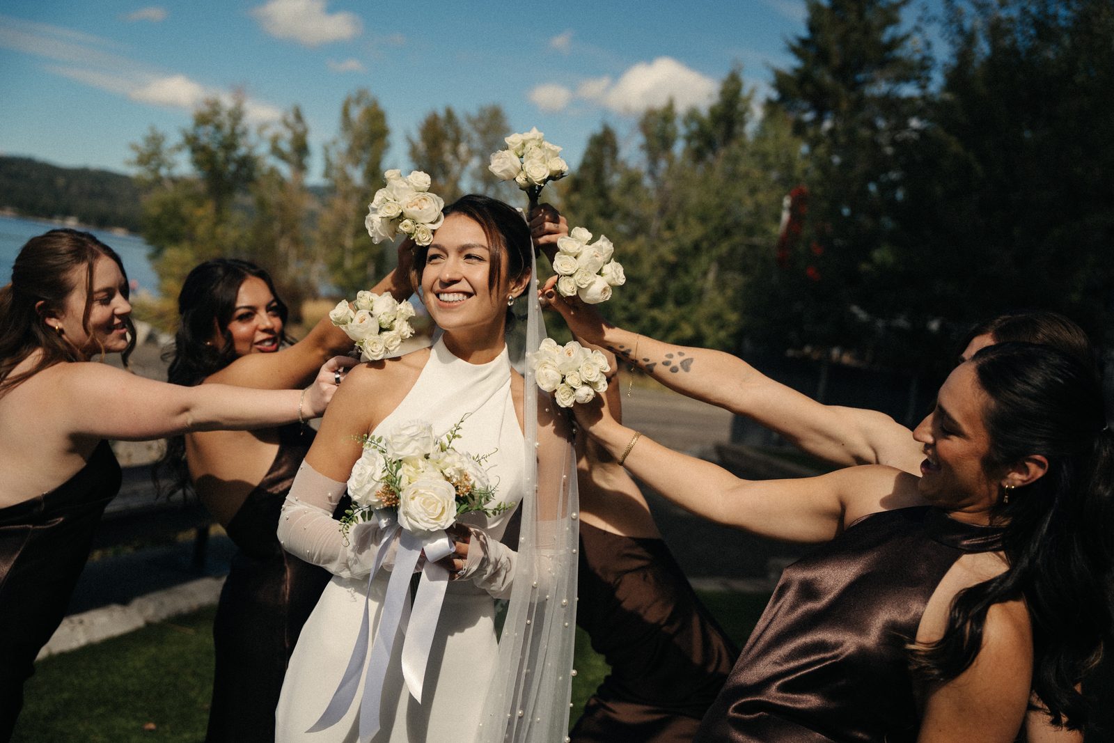 Becca laughing while her bridesmaids reach in from every side and hold white rose bouquets around her head