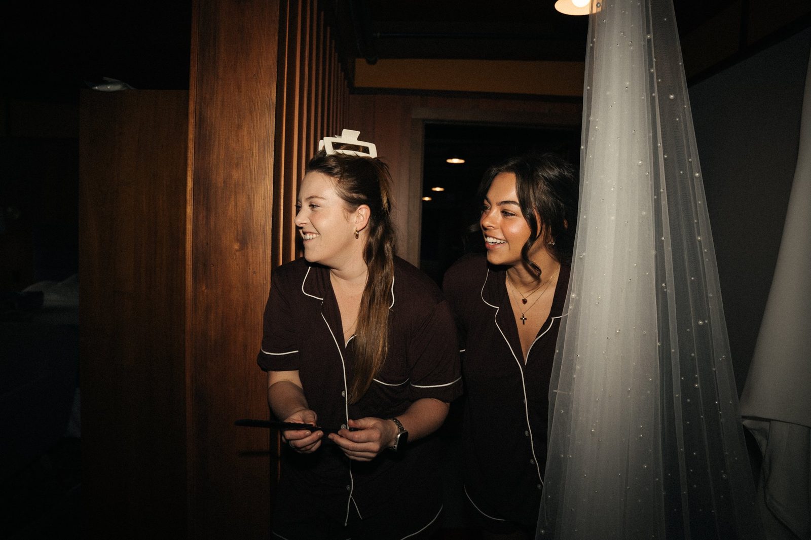 Two bridesmaids in matching brown pajamas laughing and peeking around a pearl veil in a wood-paneled hallway