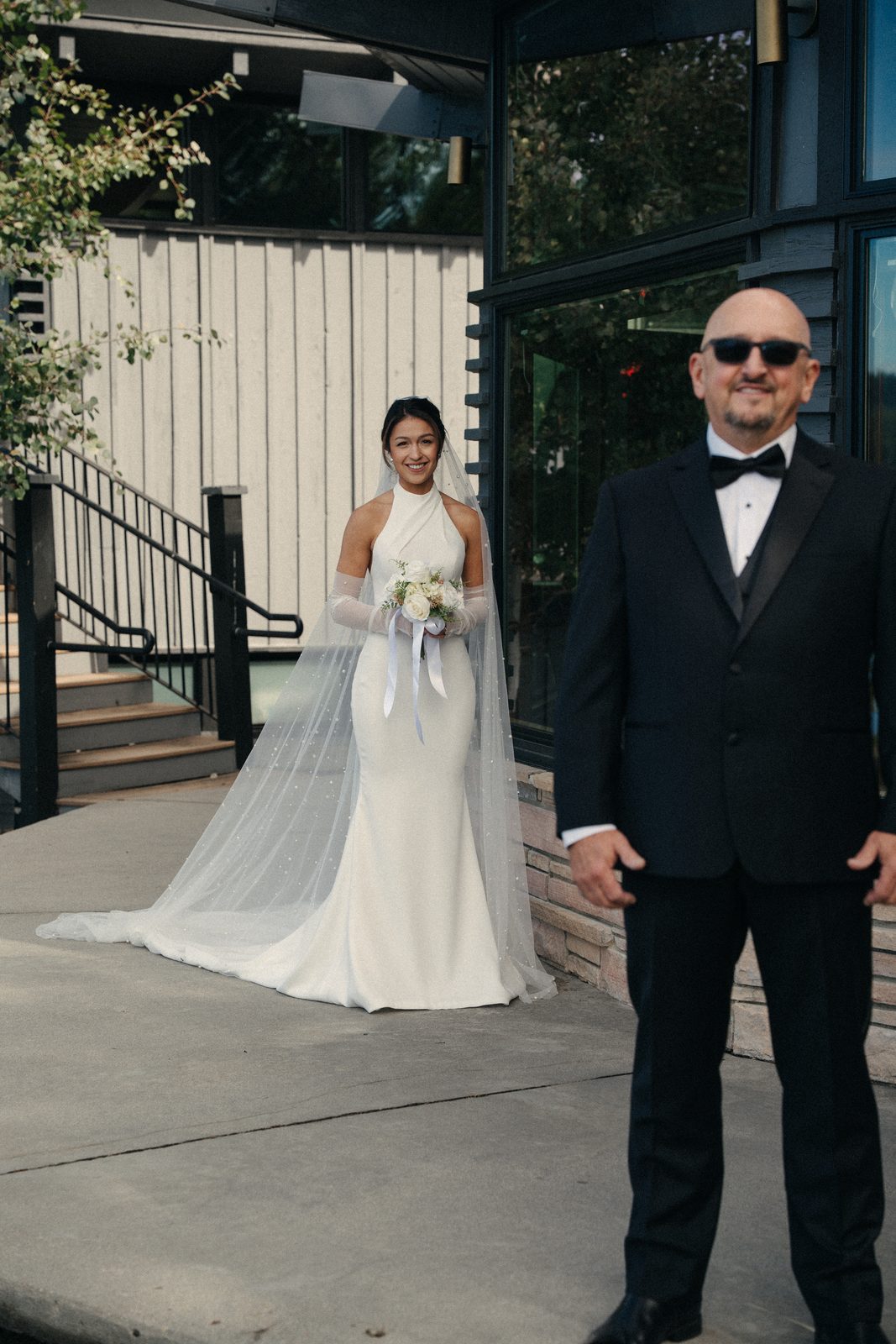 Becca’s dad stands with his back turned in sunglasses and a bow tie while Becca walks up smiling behind him for their first look