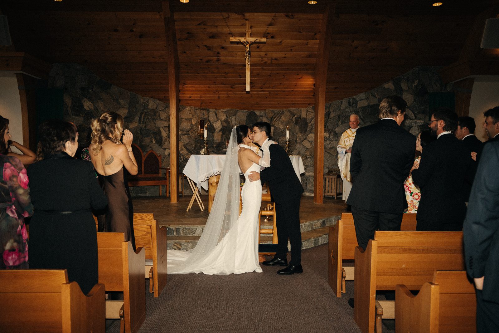 Skyler and Becca’s first kiss at the altar of a wood and stone chapel in McCall, with guests clapping and the priest smiling