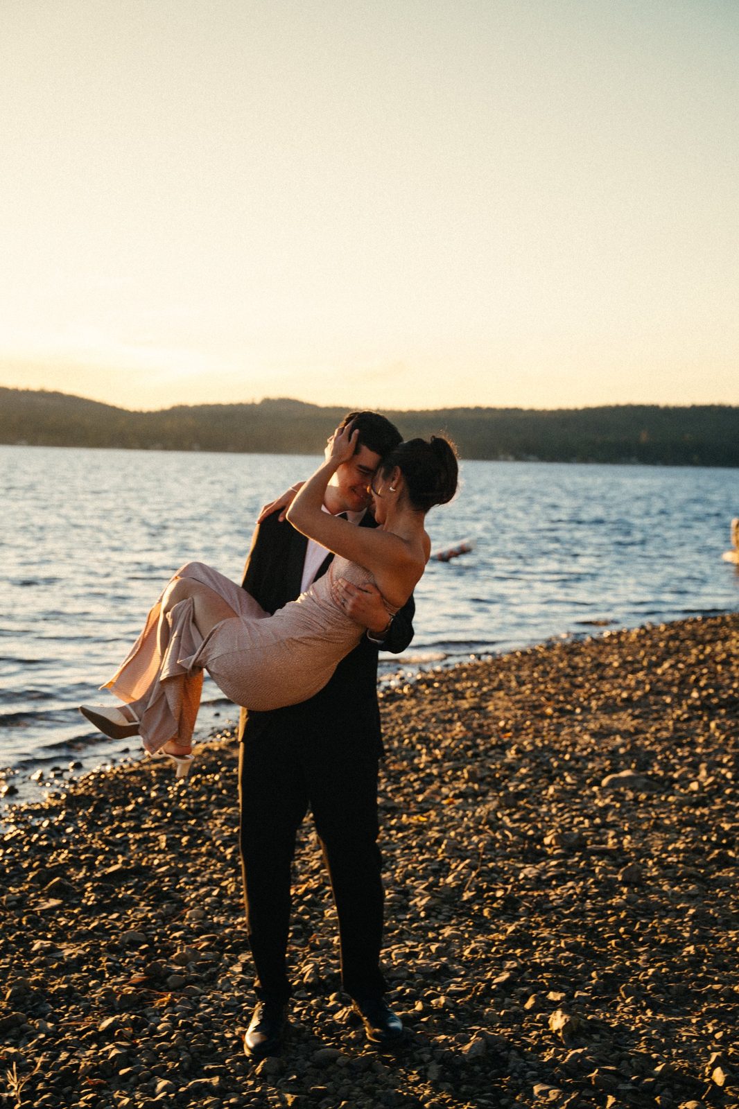 Skyler carrying Becca in his arms along the rocky lake shore at sunset, foreheads together