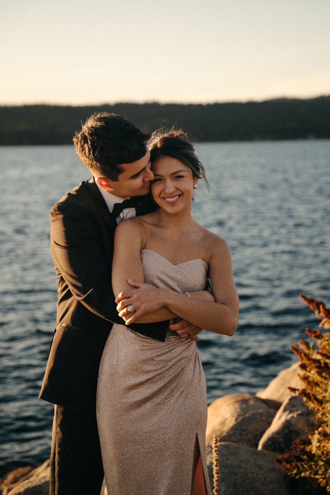 Skyler hugging Becca from behind and kissing her cheek as she smiles at the camera on the rocks by the lake at golden hour