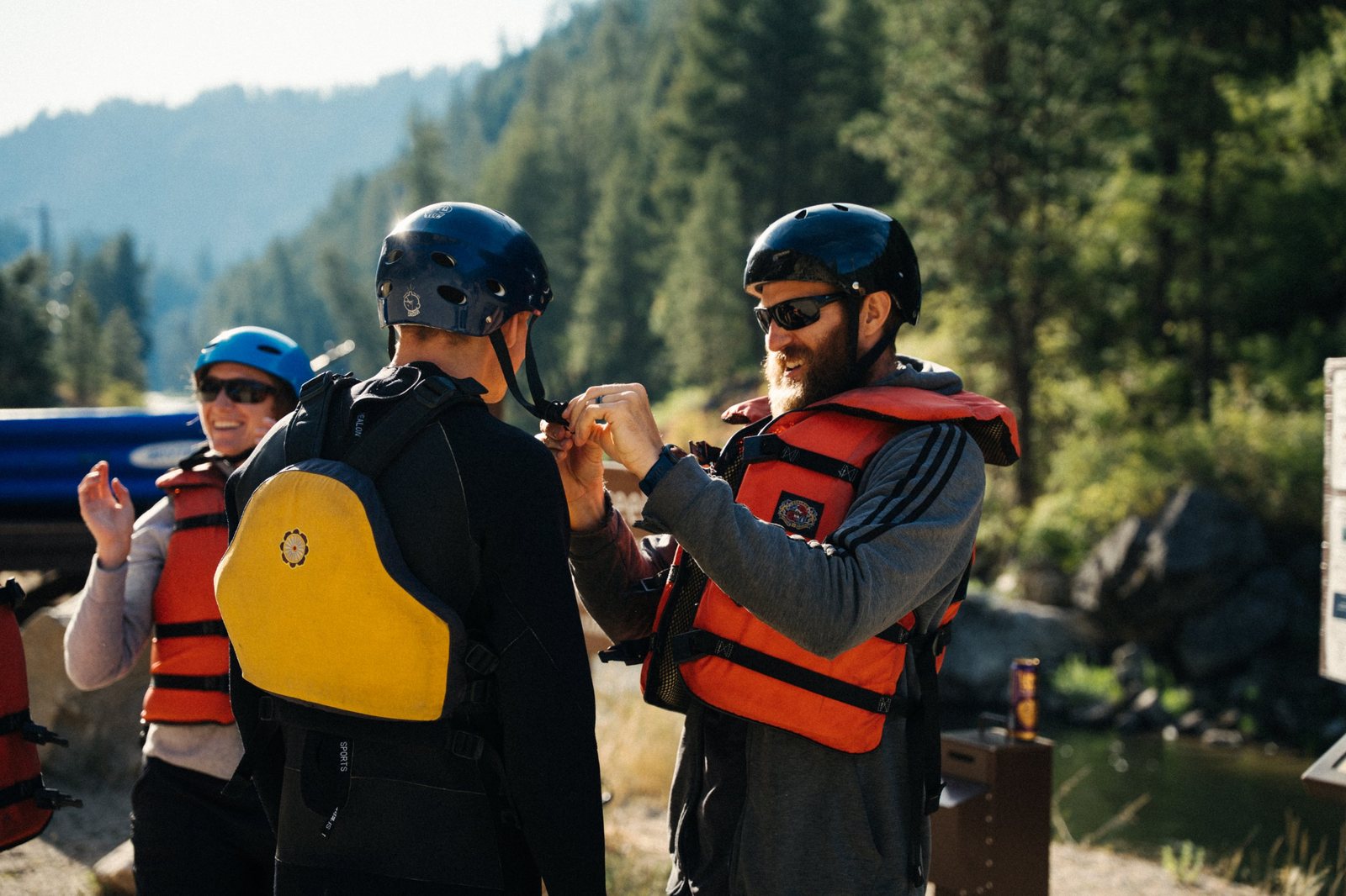 A bearded man in an orange life jacket laughing as he buckles another person's helmet strap by a river, with a woman in a blue helmet smiling behind them