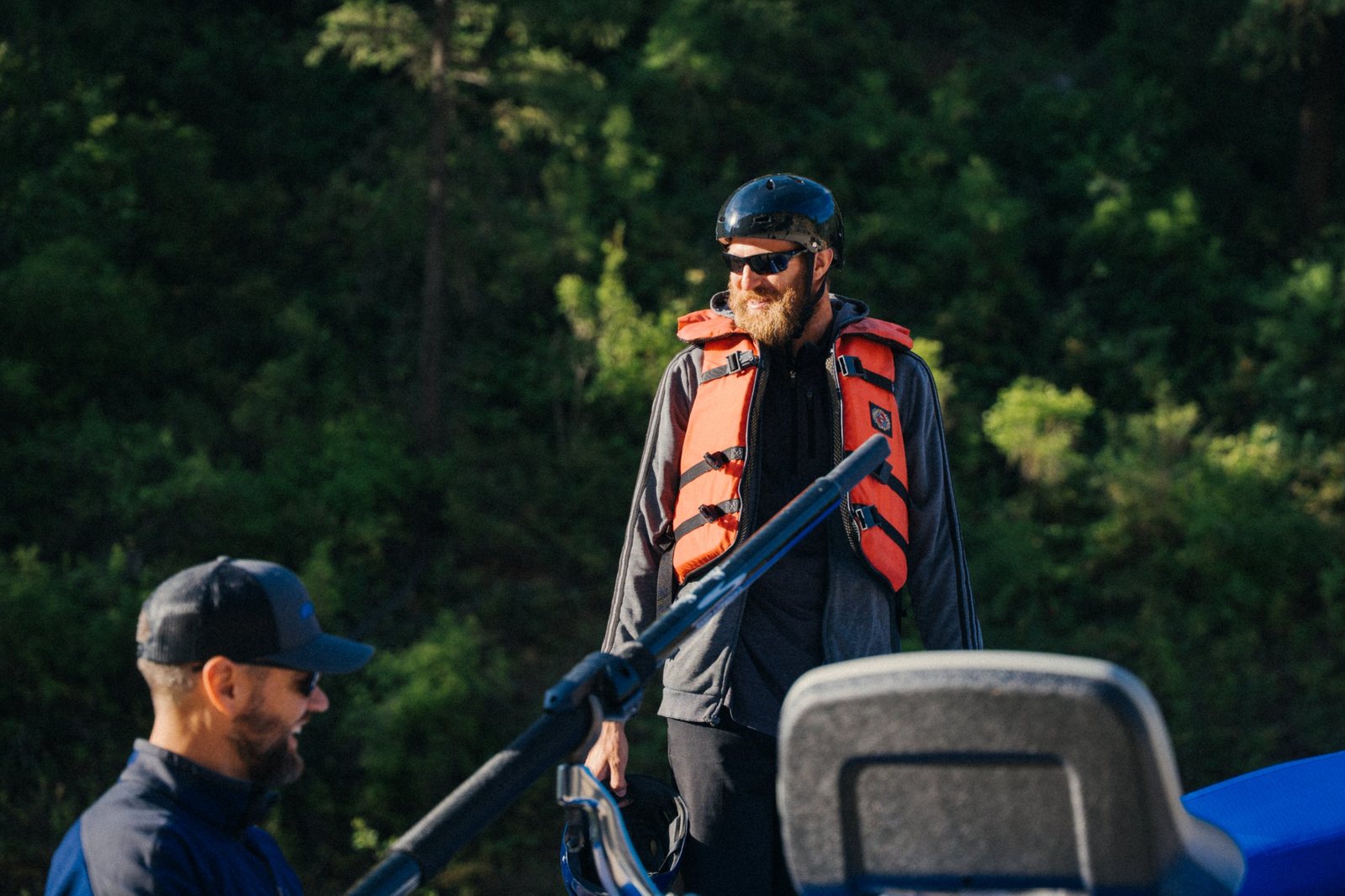 A bearded man in an orange life jacket, helmet and sunglasses smiling beside a raft, with another man laughing in the foreground and evergreens behind