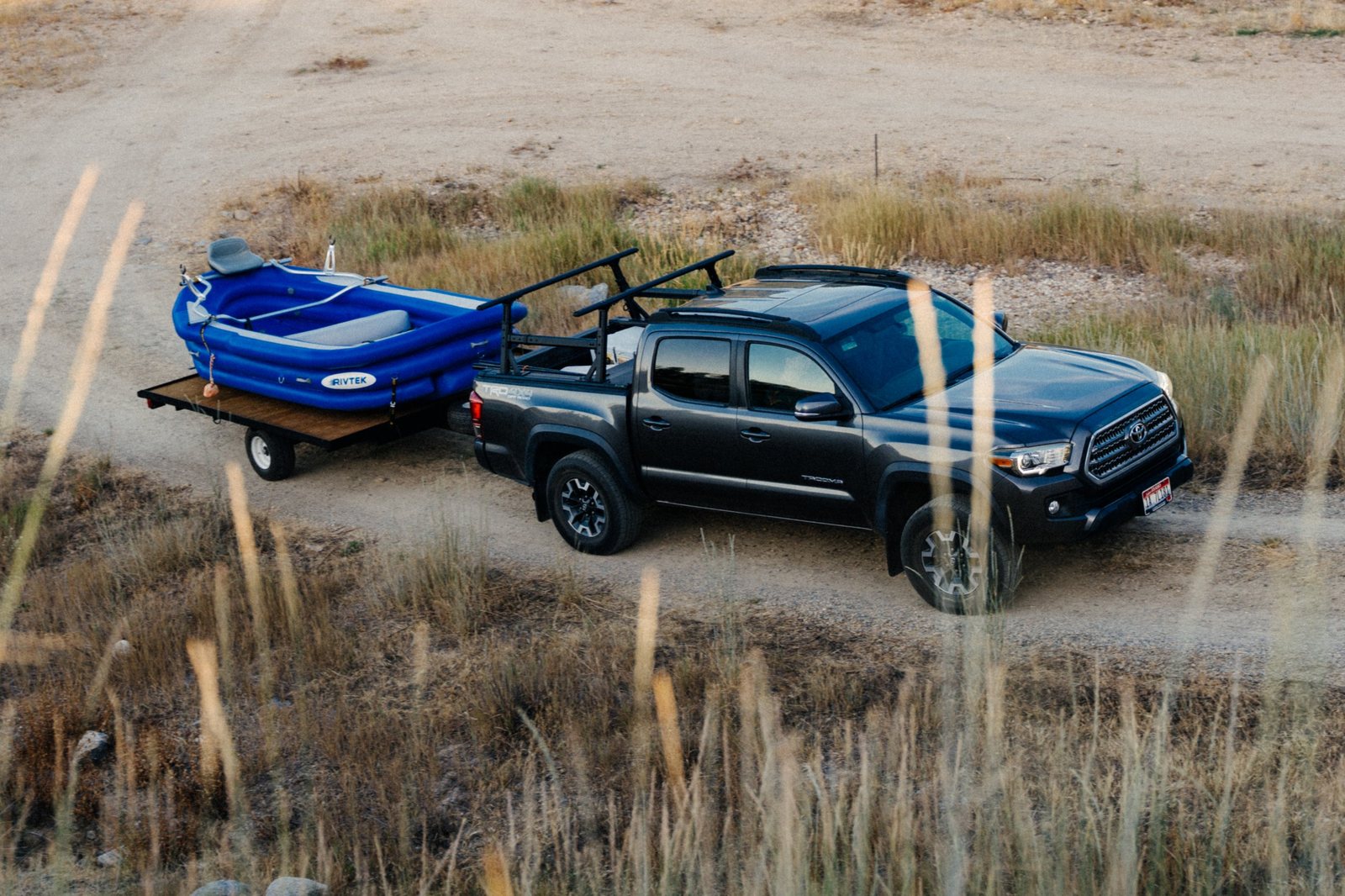 A gray pickup truck with roof racks towing a blue RIVTEK raft on a flatbed trailer down a dirt road through dry grass