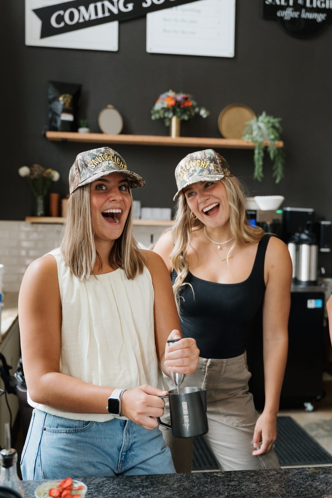 Two women in Salt & Light camo caps laughing at the bar, one using a handheld frother in a steel milk pitcher