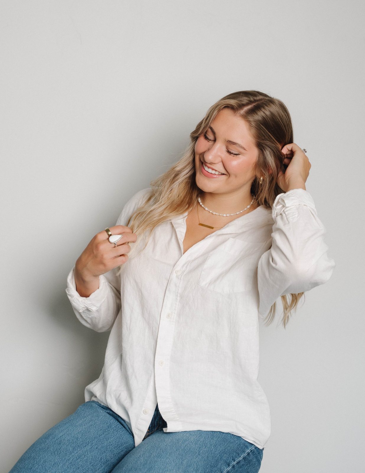 A woman in a white linen shirt looking down and smiling while tucking her hair behind her ear, against a light gray wall