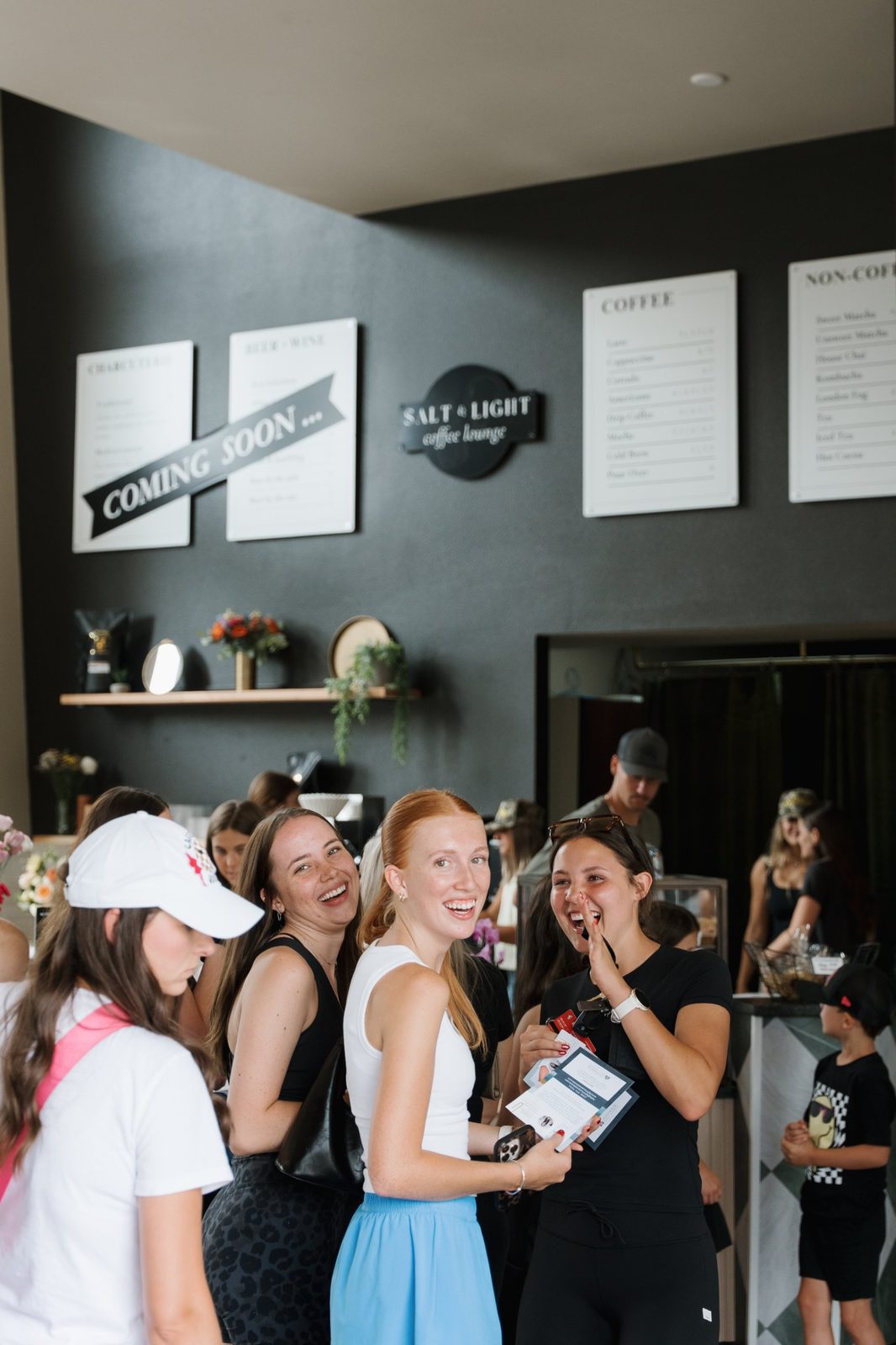 Guests laughing and looking back at the camera inside Salt & Light Coffee Lounge on grand opening day, with menu boards behind them
