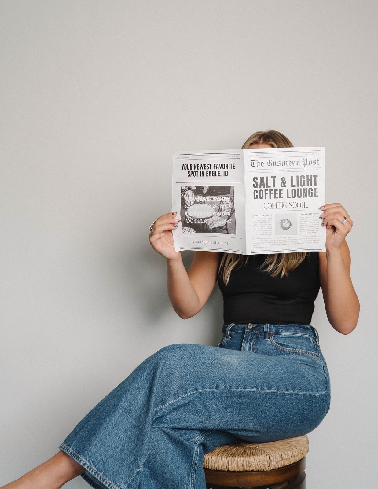 A woman in jeans sitting on a woven stool holding a Salt & Light Coffee Lounge Coming Soon newspaper in front of her face against a plain wall