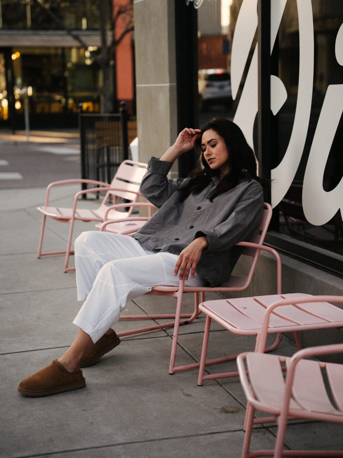 Woman in a gray linen jacket, white trousers and tan slippers lounging in a pink café chair on a downtown Boise sidewalk