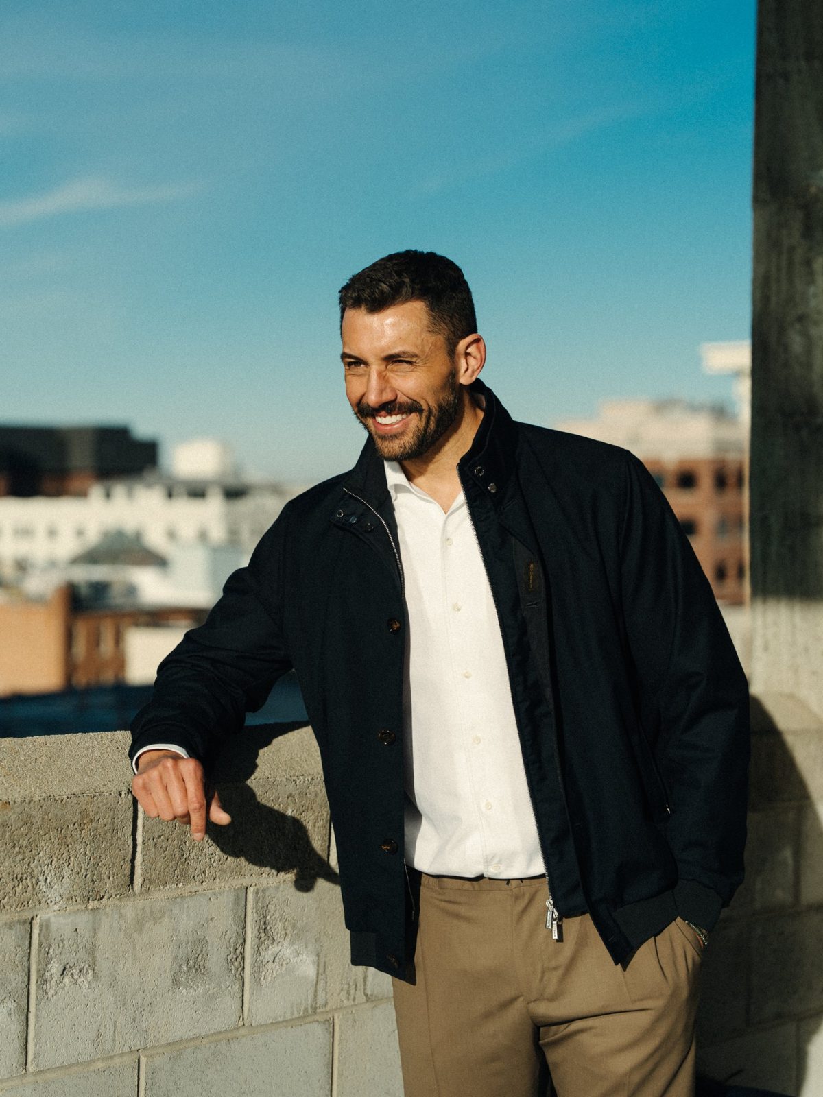 Smiling bearded man in a navy bomber jacket and white shirt leaning on a rooftop wall against a blue sky