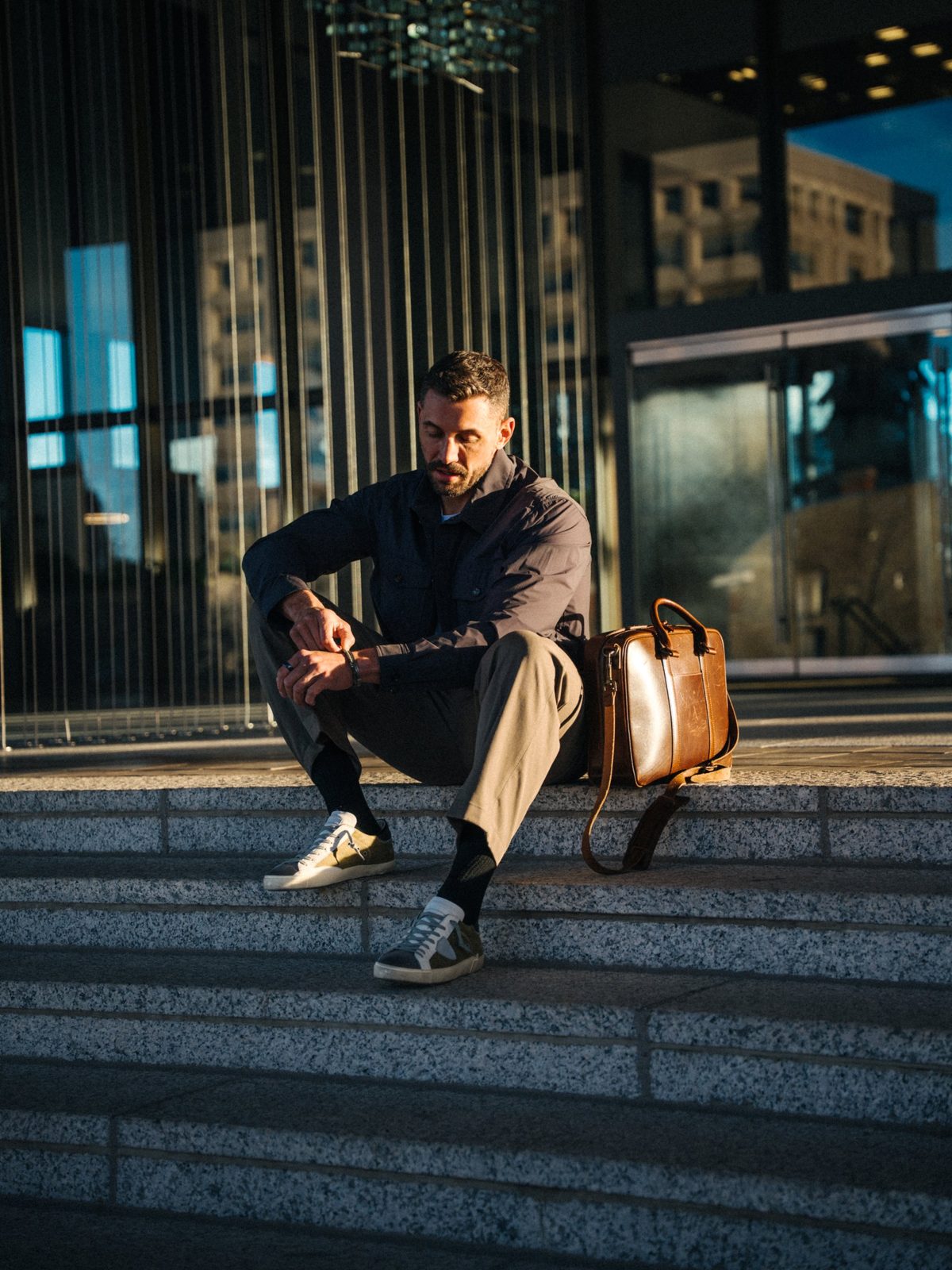 Man in a navy overshirt and tan trousers sitting on granite steps checking his watch, with a leather briefcase beside him, brand photography in Boise, Idaho