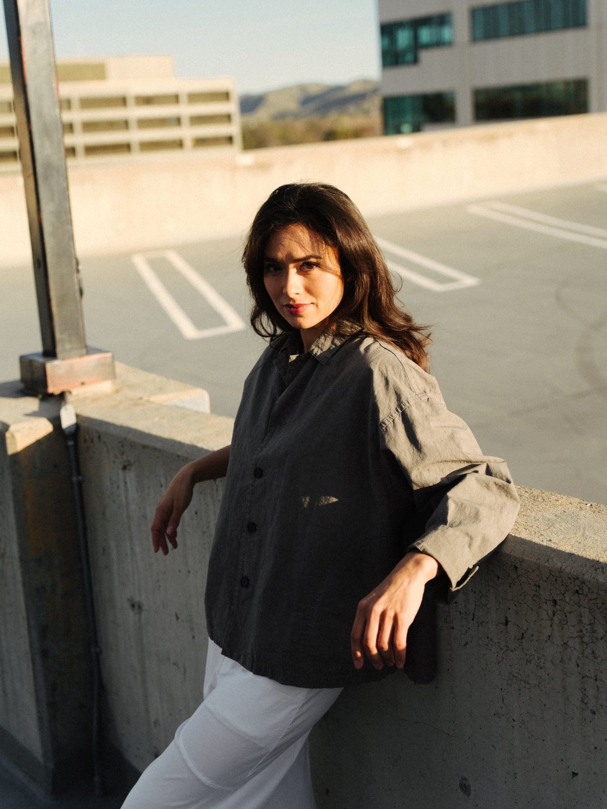 Woman in a gray linen jacket and white trousers leaning on a rooftop wall in golden light, looking toward the camera
