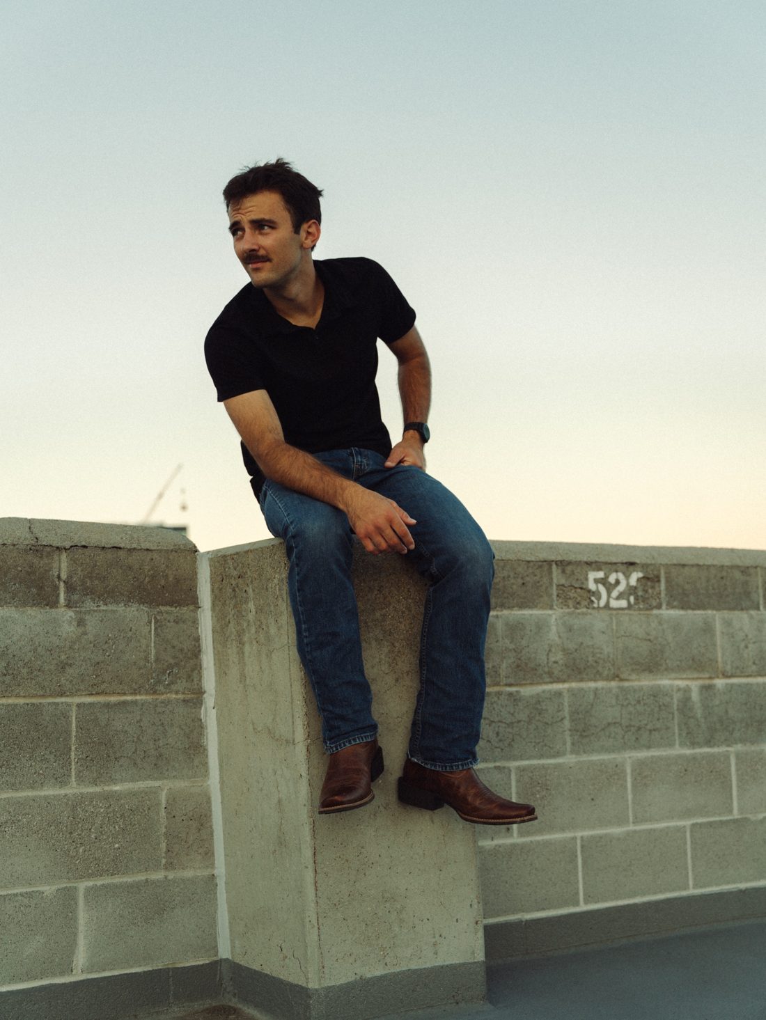 Trace Keller, Boise wedding photographer and videographer, sitting on a concrete wall at dusk in a black polo, jeans and cowboy boots