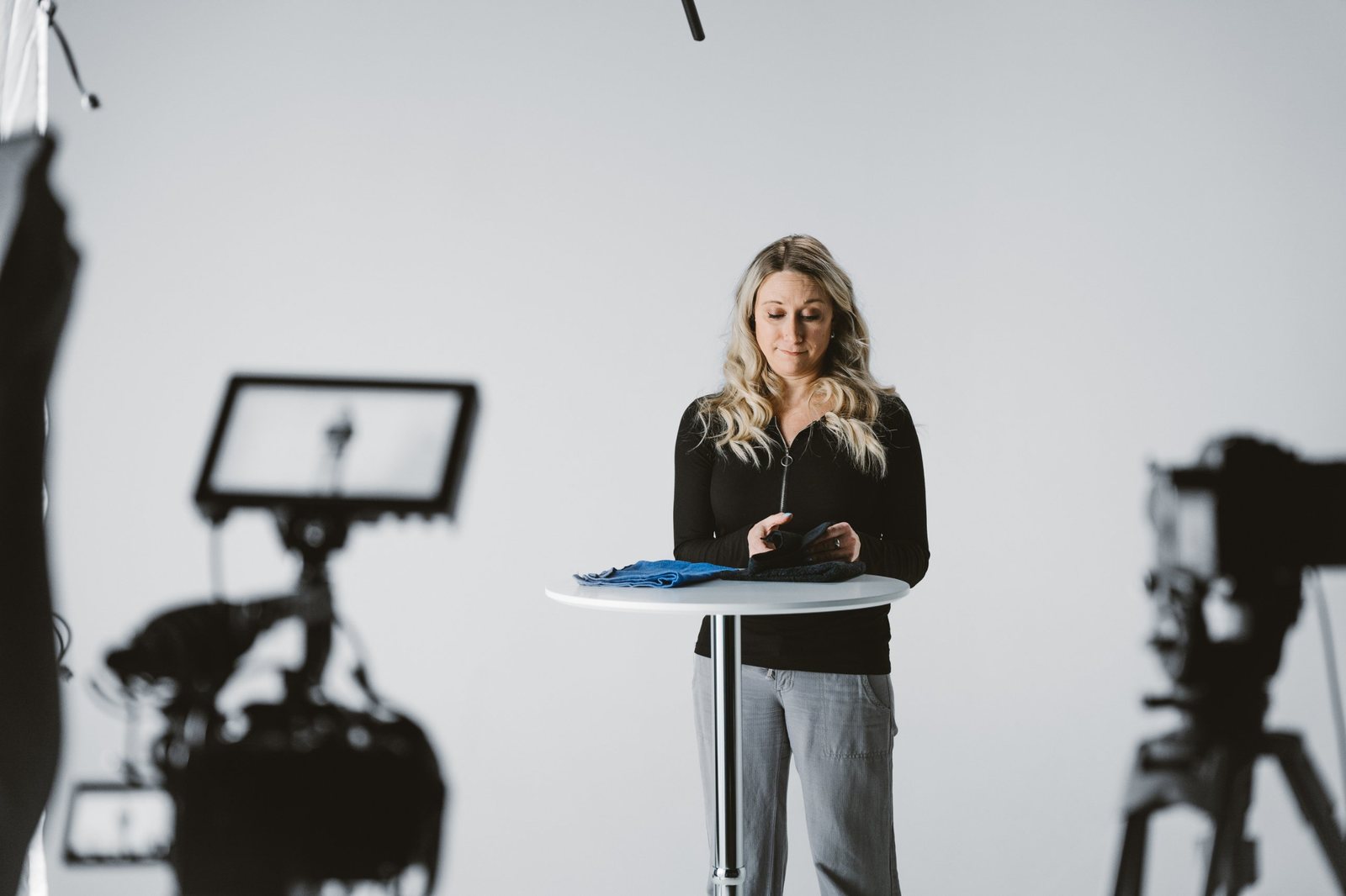 A woman in a black top looking down at a folded towel at a white standing table, with a blurred camera monitor and tripod in the foreground of the studio set